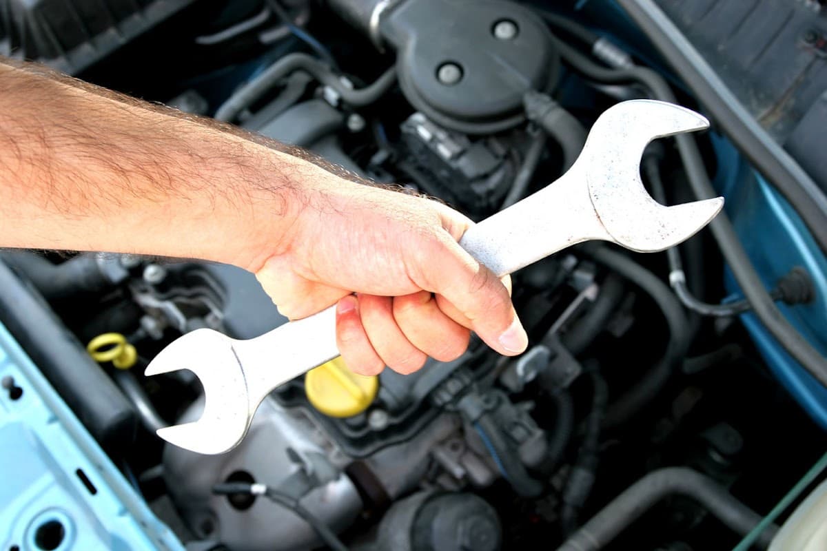 A hand holding a large wrench above an open car engine with various mechanical components visible.