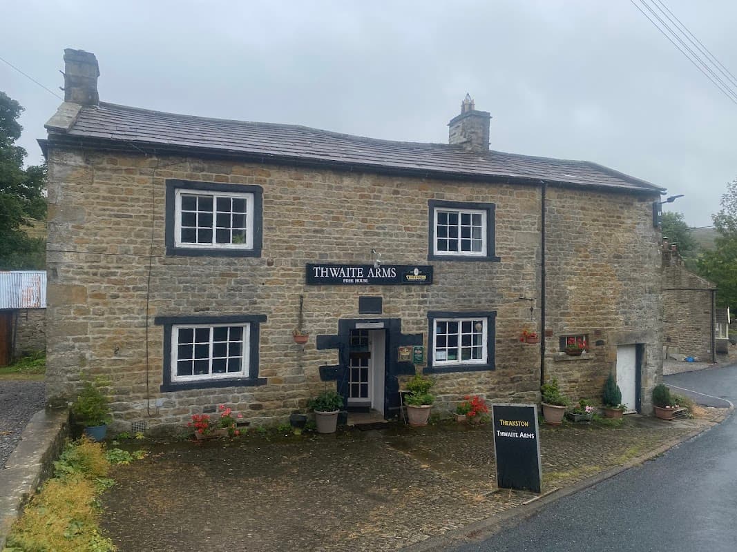 Stone building with "Thwaite Arms" sign, flower pots, and a blackboard menu, set in a rural Yorkshire landscape.