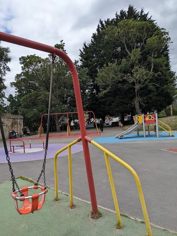 Colorful playground with swings, slides, and climbing structures, surrounded by trees and a cloudy sky.