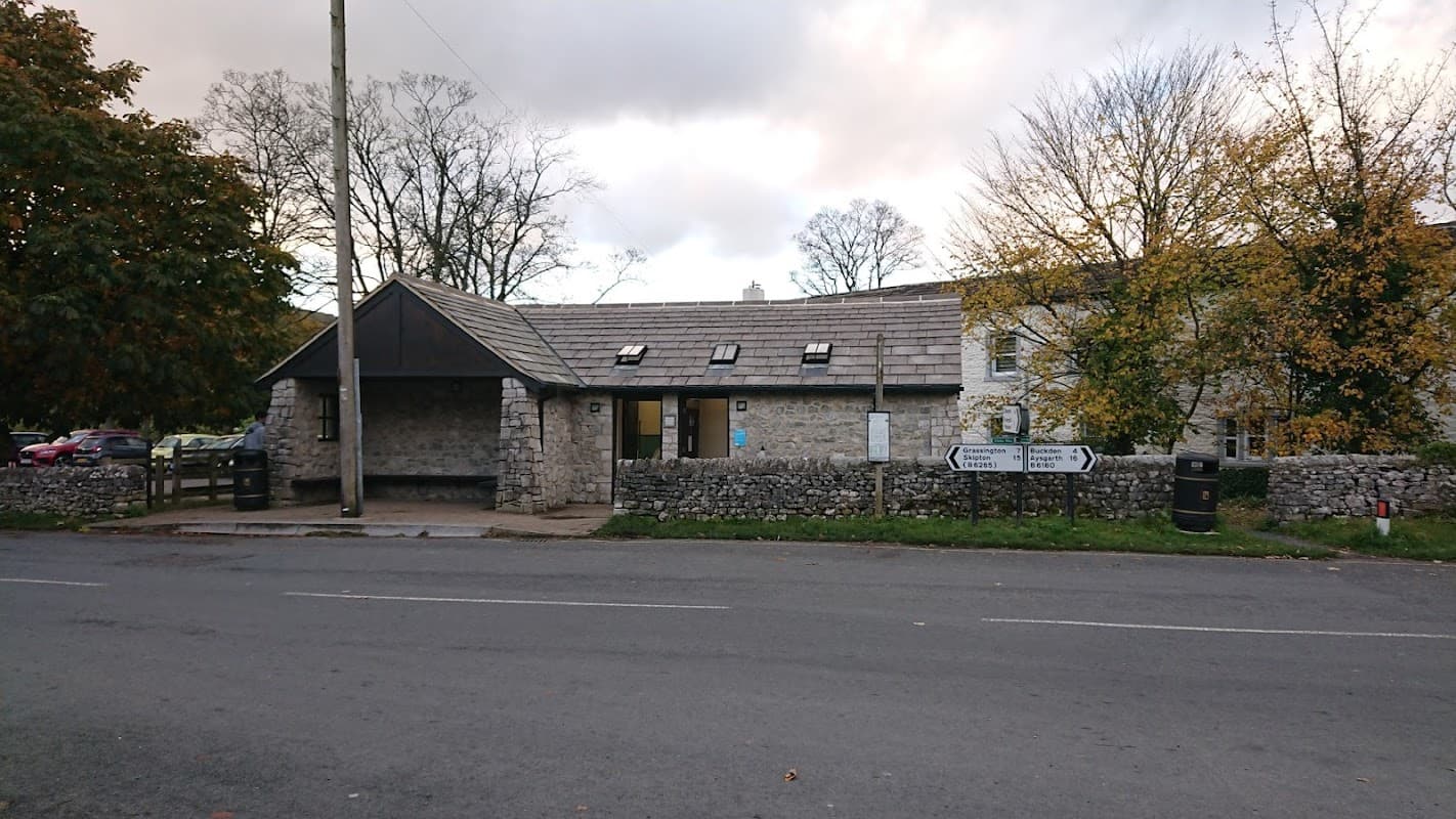 Stone building with a sloped roof, large windows, and a signpost near a quiet road surrounded by trees in Kettlewell.
