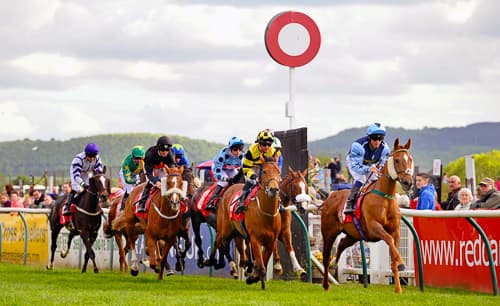 Horses racing at Redcar Racecourse, with colorful jockeys and a large red and white marker in the background.