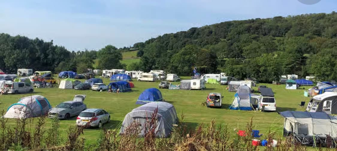 Camping site with numerous tents and caravans set in a green landscape, surrounded by trees under a clear blue sky.