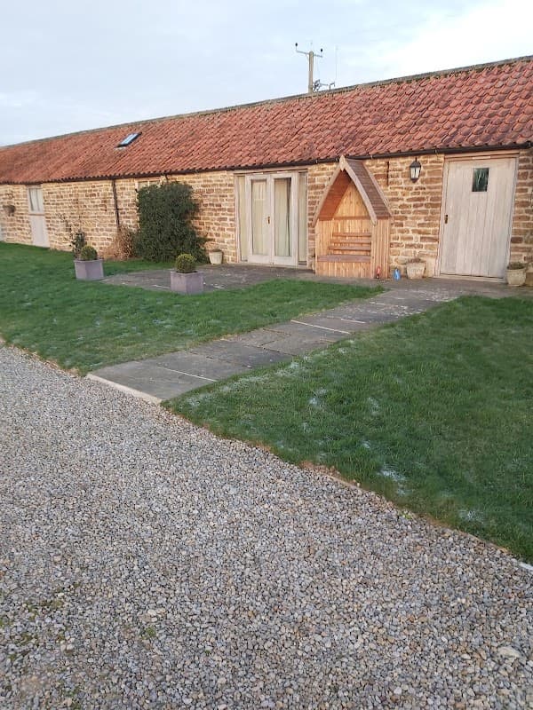 Stone cottage with a red-tiled roof, wooden doors, and a gravel pathway surrounded by grass and potted plants.