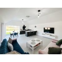 Modern living room with blue and pink sofas, white coffee table, wall-mounted TV, and open kitchen with black dining set.