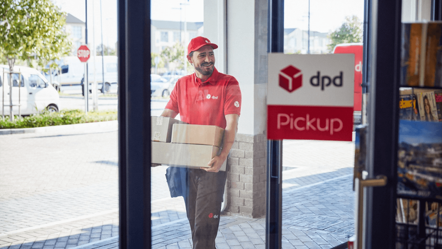 A delivery person in a red uniform carries a package through the entrance of a DPD Pickup Parcelshop.