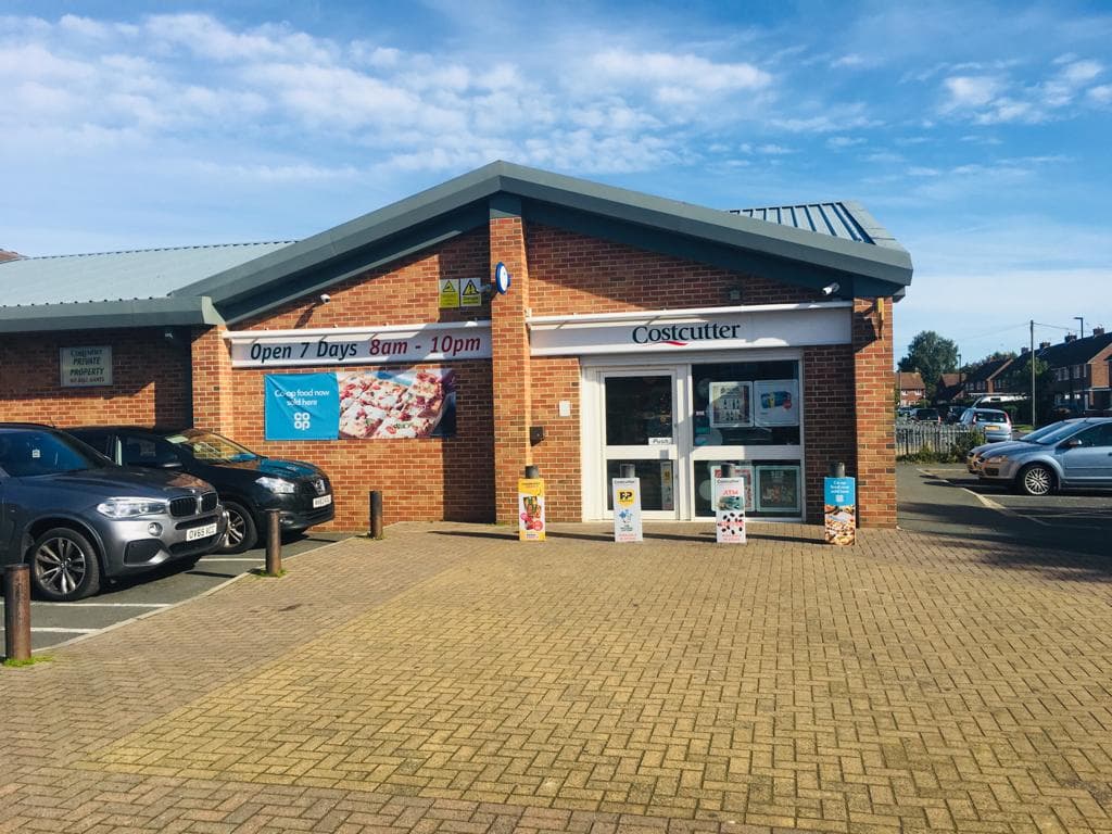 Costcutter store with a blue banner, open 7 days, surrounded by parked cars and a clear blue sky.