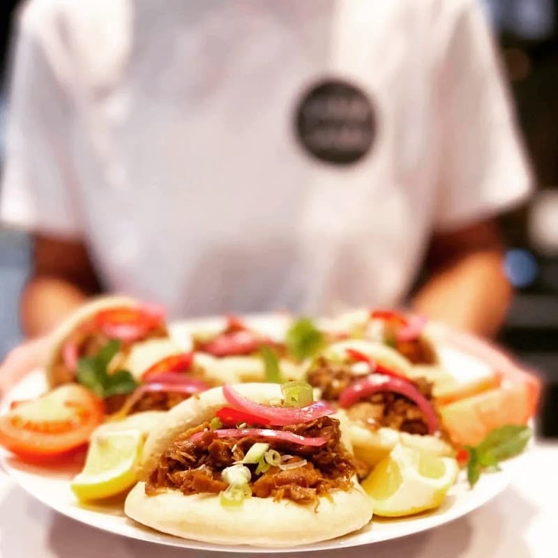 A plate of bao buns filled with shredded meat, garnished with pickled onions, lime wedges, and fresh herbs.