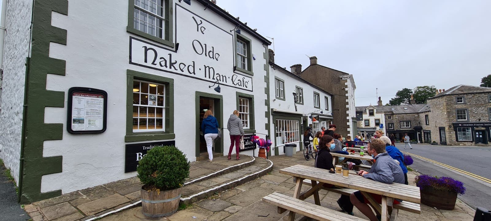 Historic building with "Ye Olde Naked Man Café" sign, outdoor seating, and people enjoying drinks in Settle, Yorkshire.