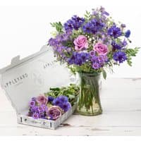 Bouquet of purple and pink flowers in a glass vase, with matching blooms in an open Appleyard box nearby.